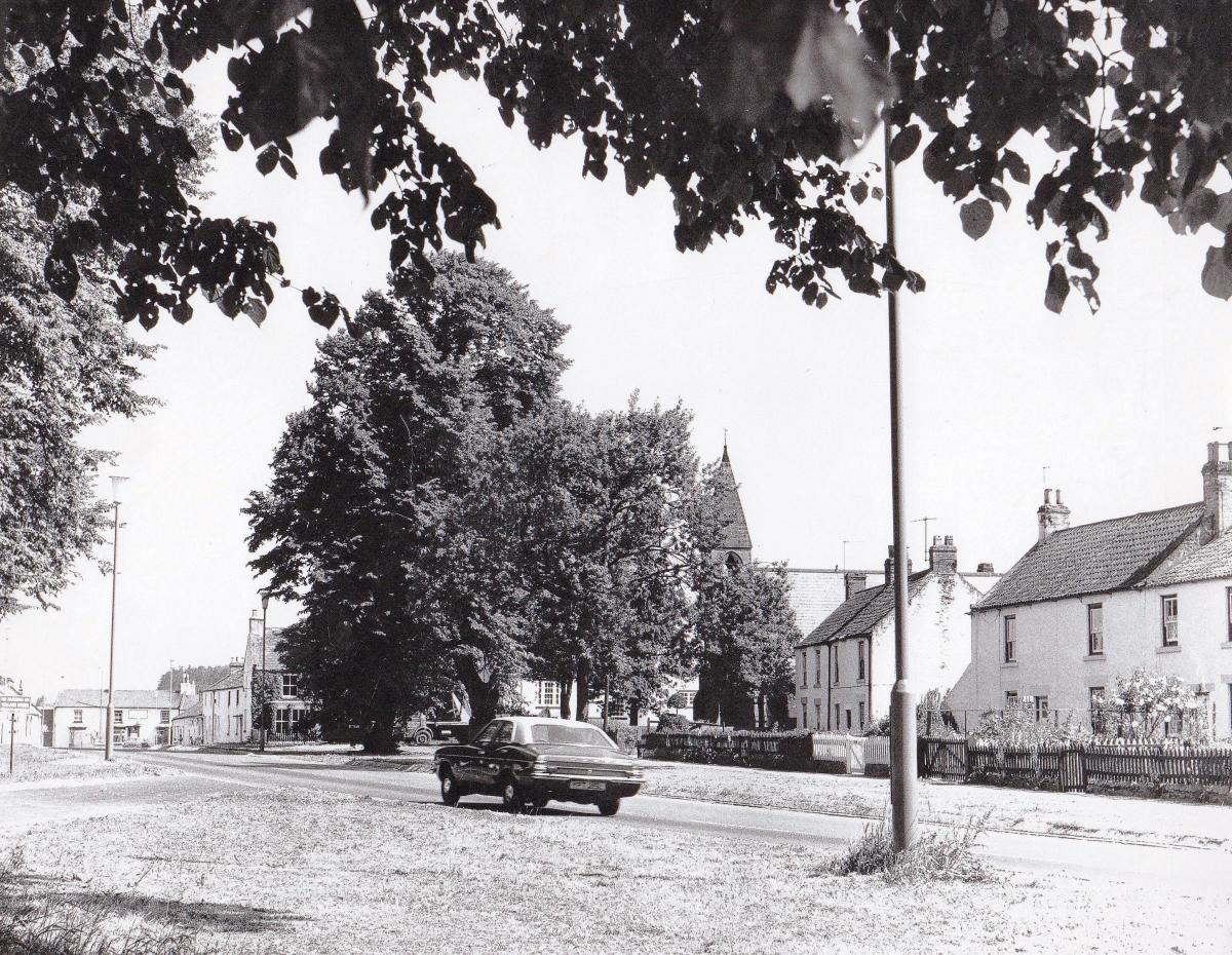 A view from the village green, taken in 1976. St Mary’s church can be seen behind the trees as a Mark III Ford Cortina races up the B6275