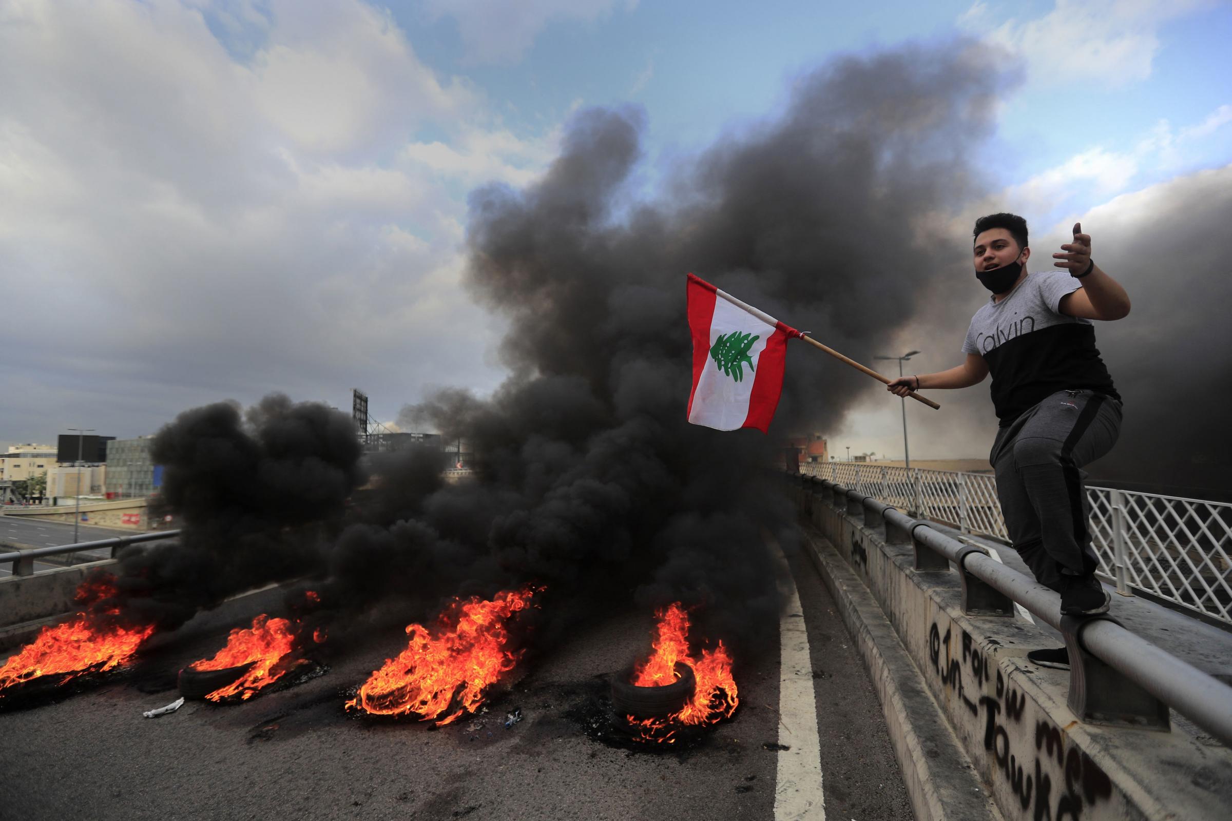 A protestor waves a Lebanese flag near burning tires set to block a main highway, during a protest in the town of Jal el-Dib, north of Beirut, Lebanon Picture: HUSSEIN MALLA/AP
