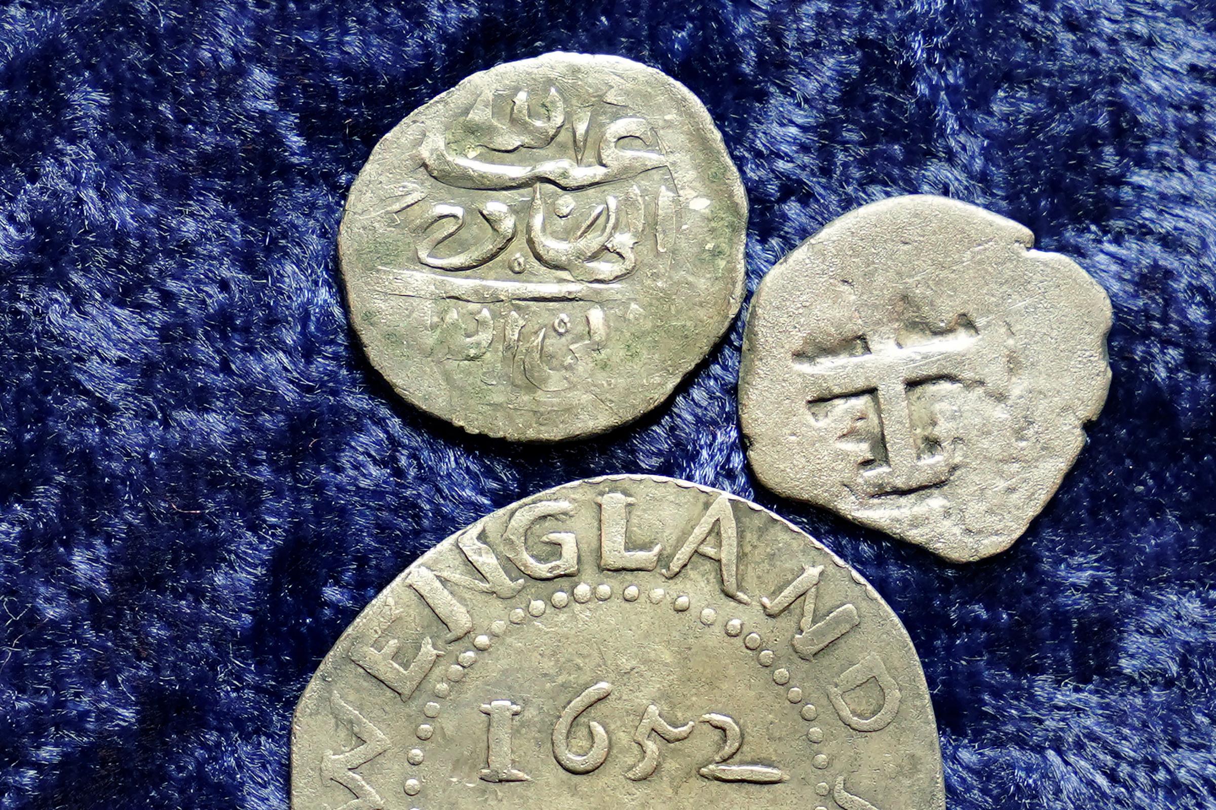 A 17th century Arabian silver coin, top, that research shows was struck in 1693 in Yemen, rests near an Oak Tree Shilling minted in 1652 by the Massachusetts Bay Colony, below, and a Spanish half real coin from 1727, right. The Arabian coin was found at