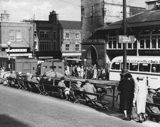 A busy High Row in 1964: there are large numbers of older people sitting on the benches in the sun or standing in the queues beneath the clock tower. Practically all of them are wearing some form of headgear
