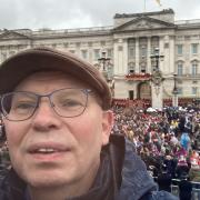Chris Lloyd on the Victoria Monument with King Charles and Queen Camilla waving at the camera from the balcony behind